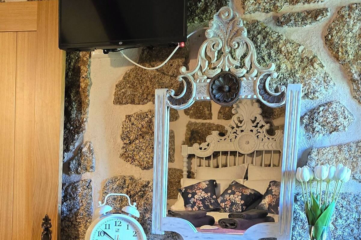 A stone wall with a mounted television, a decorative mirror, a clock, and a small table with a vase of flowers.