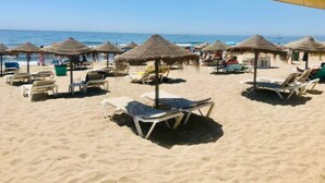 Una playa con tumbonas y sombrillas de paja, cielo despejado y el océano al fondo.