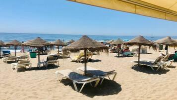 A beach with sun loungers and thatched umbrellas, a clear sky, and the ocean in the background.