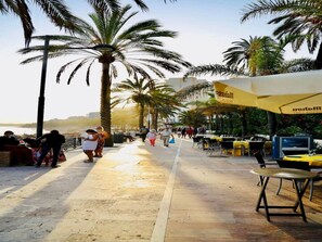 Une promenade en bord de mer, bordée de palmiers, avec des espaces de repos en plein air et une vue sur la mer.