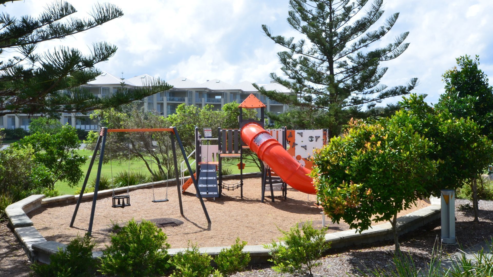 A playground with swings and a slide, surrounded by trees and a building in the background.