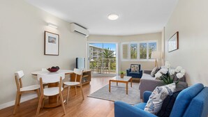 A modern living room with a dining area, a sofa, a TV, and a balcony with a view of the building. - Resort on the Beach 5114 15 with resort Pool (Kingscliff)