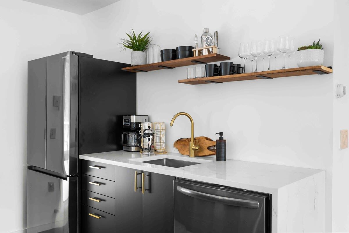 A modern kitchen with a stainless steel refrigerator, a coffee machine, a sink with a gold faucet, and wooden shelves above holding various kitchen items.