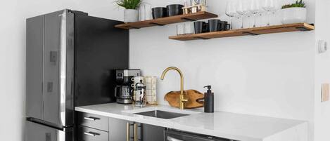 A modern kitchen with a stainless steel refrigerator, a coffee machine, a sink with a gold faucet, and wooden shelves above holding various kitchen items.