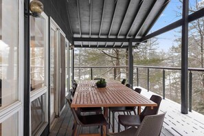 Un espacio comedor al aire libre cubierto, con mesa y sillas de madera, ventanas amplias y vistas a árboles.