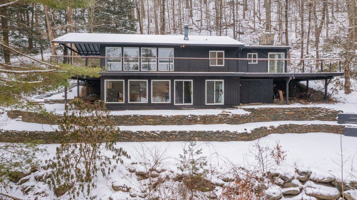 Une maison moderne à deux étages, dotée de grandes fenêtres et d’une fondation en pierre, entourée d’un paysage enneigé et d’arbres.