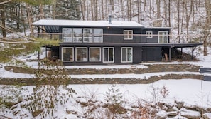 Une maison moderne à deux étages, dotée de grandes fenêtres et d’une fondation en pierre, entourée d’un paysage enneigé et d’arbres.