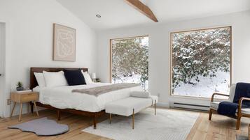 A bedroom with a large bed, a bedside table with a lamp, a chair, and a view of snow-covered trees through large windows.