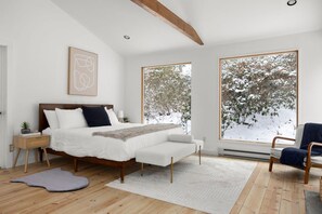 Un dormitorio con una cama grande, una mesita de noche con lámpara, una silla y vistas a árboles cubiertos de nieve a través de amplias ventanas.