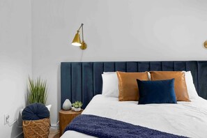 A neatly made bed with a dark headboard, a bedside lamp, and a small wooden nightstand with a plant and decorative items.
