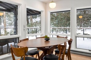 Uma área de refeições com uma mesa redonda e cadeiras, janelas amplas com vista para uma paisagem nevada e uma luminária pendente sobre a mesa.