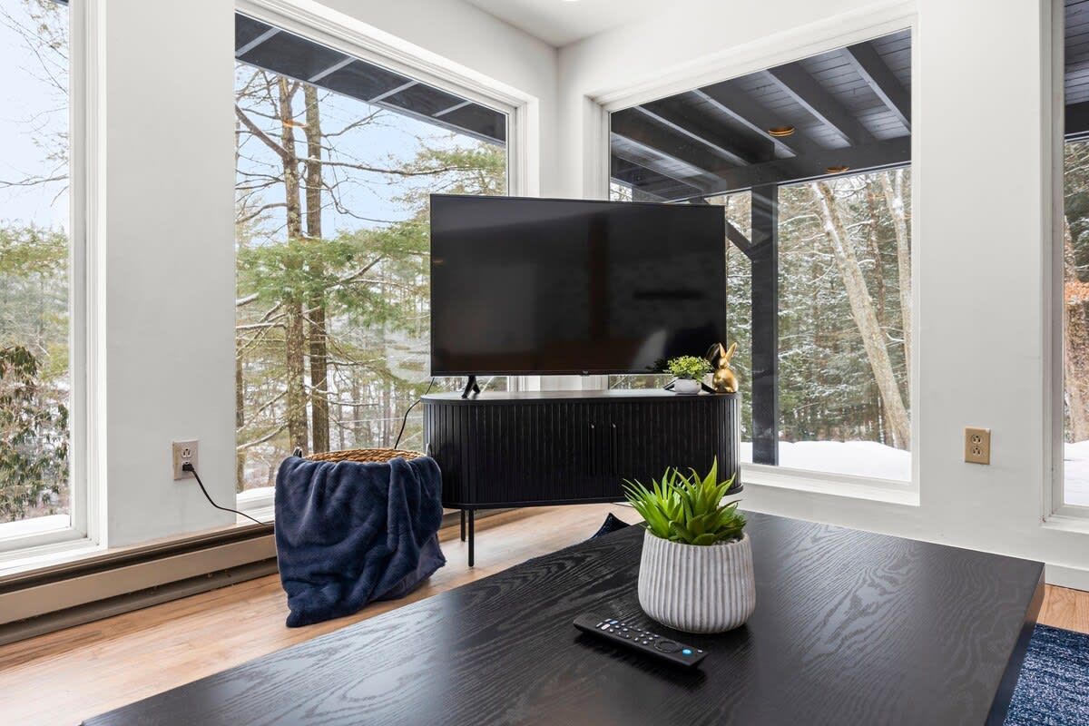 Een moderne woonkamer met een grote flatscreen televisie bevestigd aan een zwart kastje, een houten salontafel met een afstandsbediening en een bloempot.