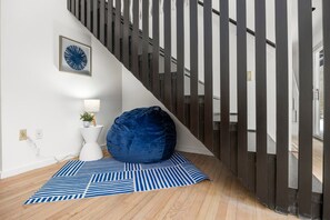 A modern interior with a wooden floor, a blue and white striped rug, a dark blue bean bag, a white side table with a lamp, and a framed artwork on the wall.