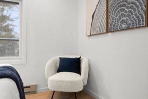A room with a white chair, a window, and framed artwork on the wall.
