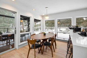 Un comedor moderno con una mesa redonda y sillas, ventanas amplias y una zona de cocina con taburetes de bar.