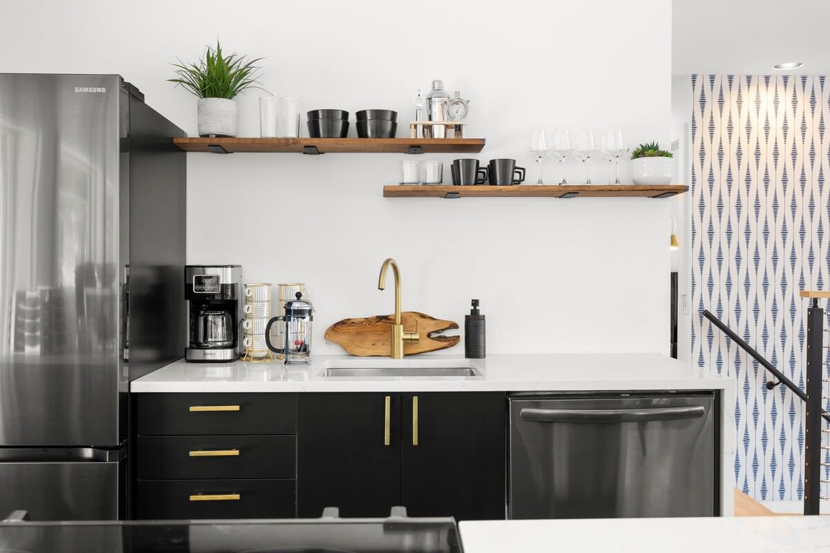 A modern kitchen with a stainless steel refrigerator, black cabinets, gold handles, a coffee maker, and wooden shelves with glassware.