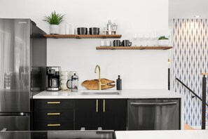 A modern kitchen with a stainless steel refrigerator, black cabinets, gold handles, a coffee maker, and wooden shelves with glassware.