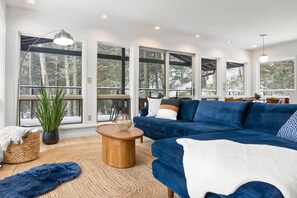 A modern living room with a blue sofa, a wooden coffee table, and large windows offering a view of trees.
