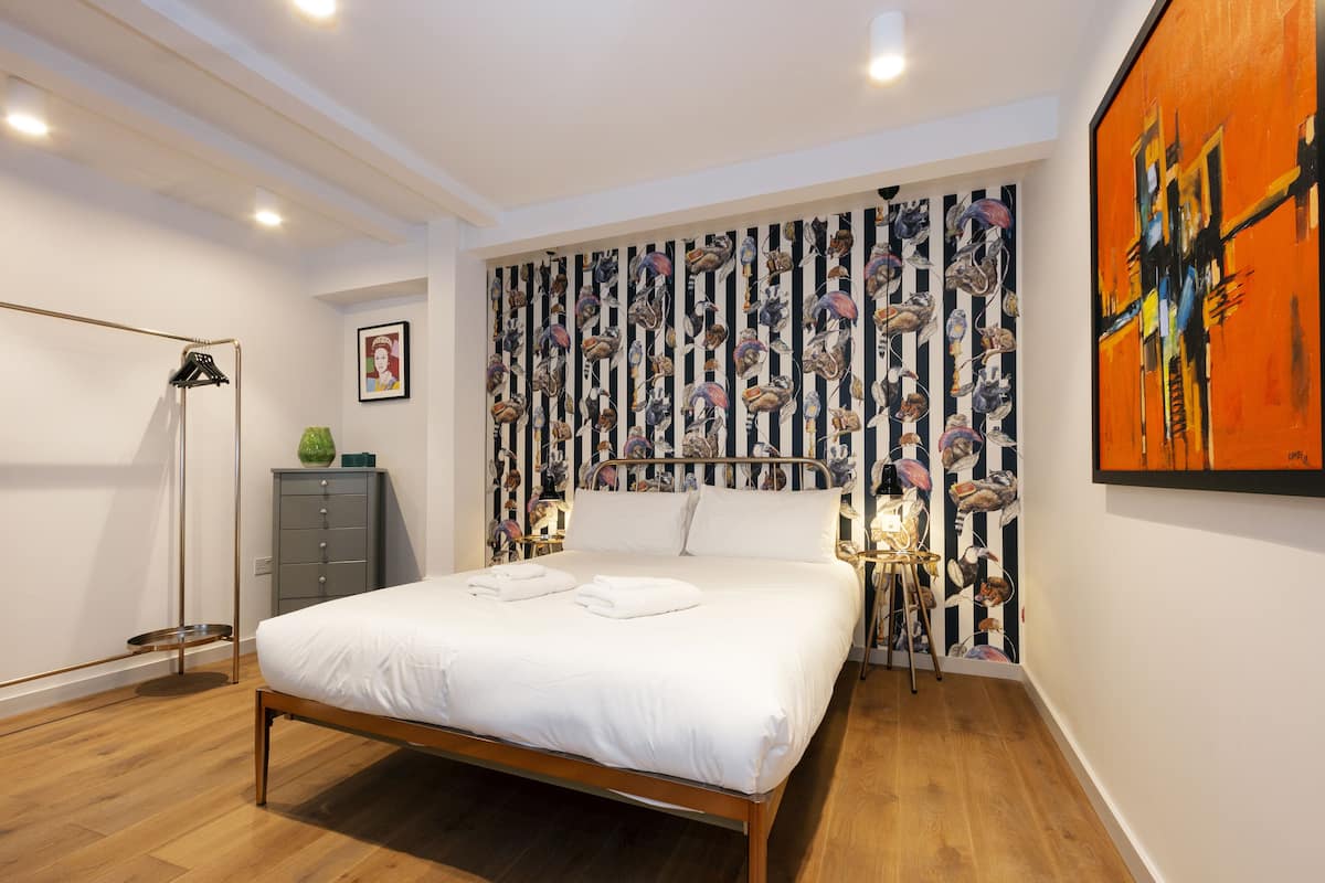 A bedroom with a bed, a wardrobe, a floor lamp, a painting, and a patterned wallpaper.