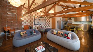 Un salón espacioso con dos sofás informales de color gris, vigas de madera, una pared de ladrillo y una cocina al fondo.