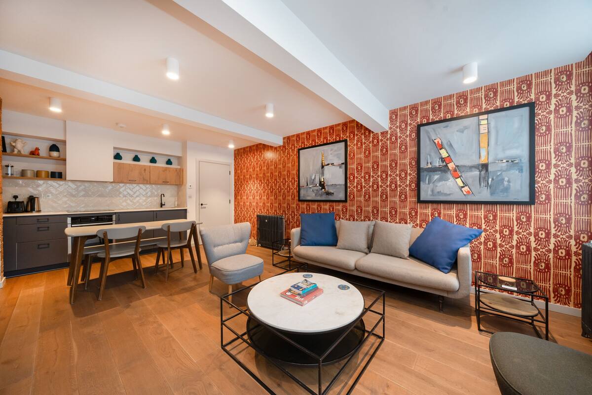 A modern living room with a sofa, coffee table, and chairs. There is a kitchen area with cabinets and a countertop.