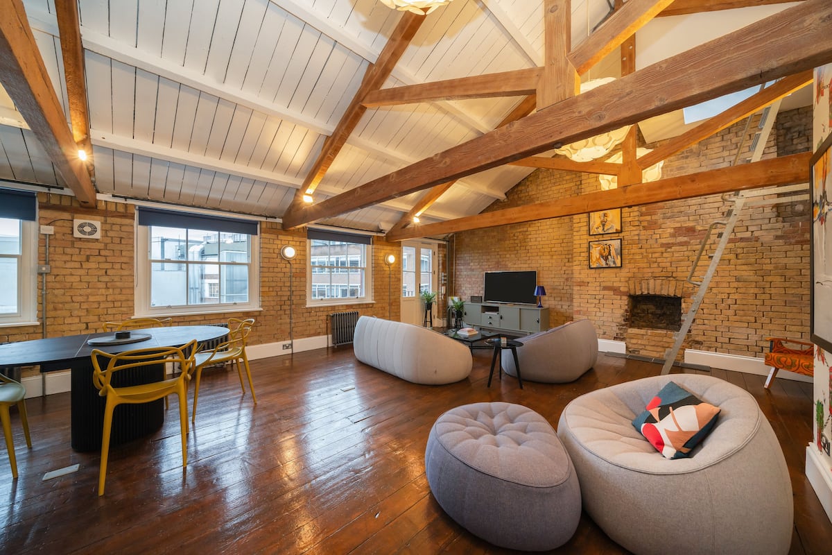 A spacious loft with exposed wooden beams, a brick wall, a fireplace, and a large window.