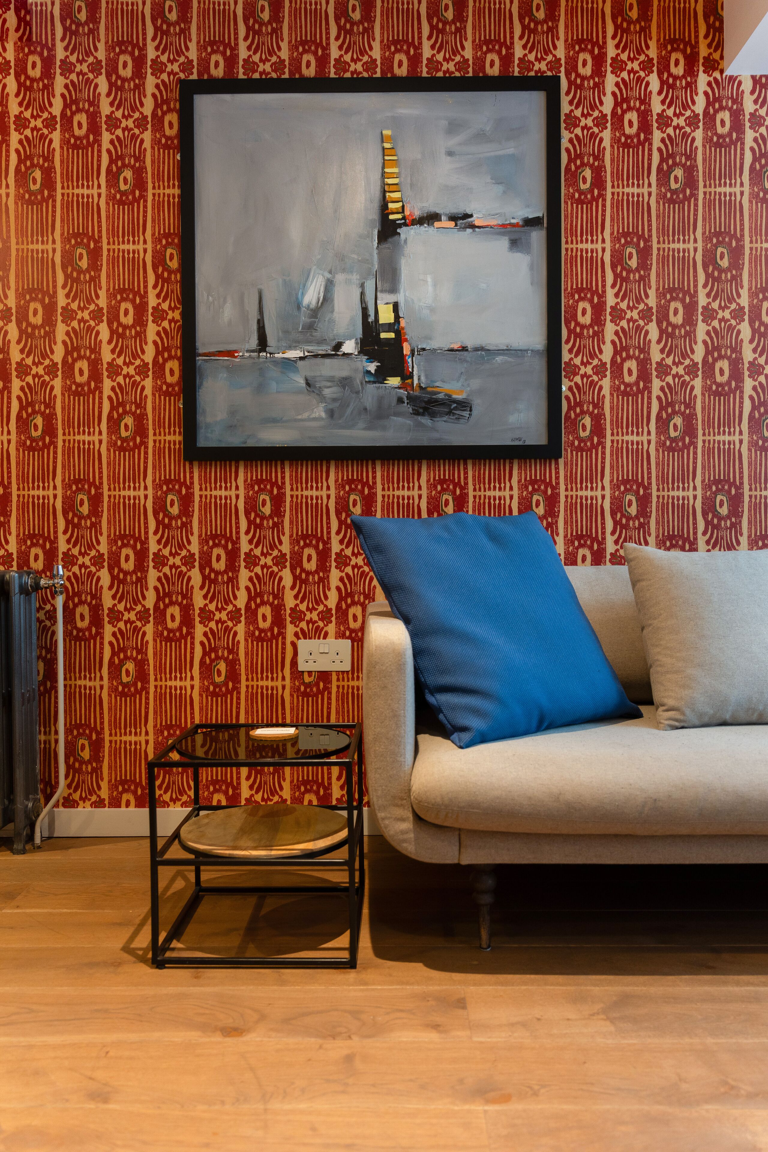 A room with a patterned wallpaper, a framed abstract painting, a beige sofa with a blue pillow, and a small black table with a round glass top.