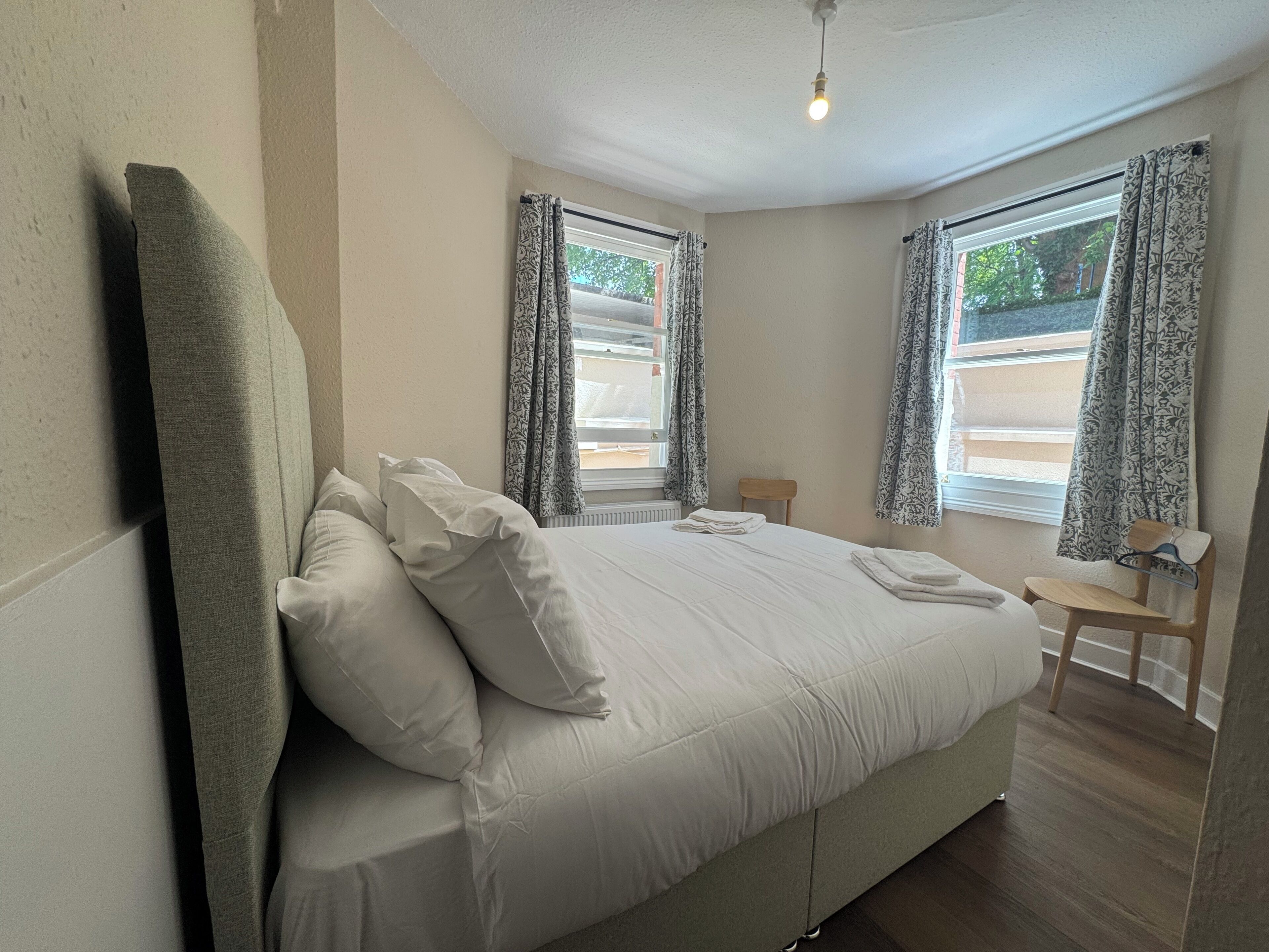 A bedroom with a large bed, two windows with patterned curtains, a wooden chair, and a radiator.