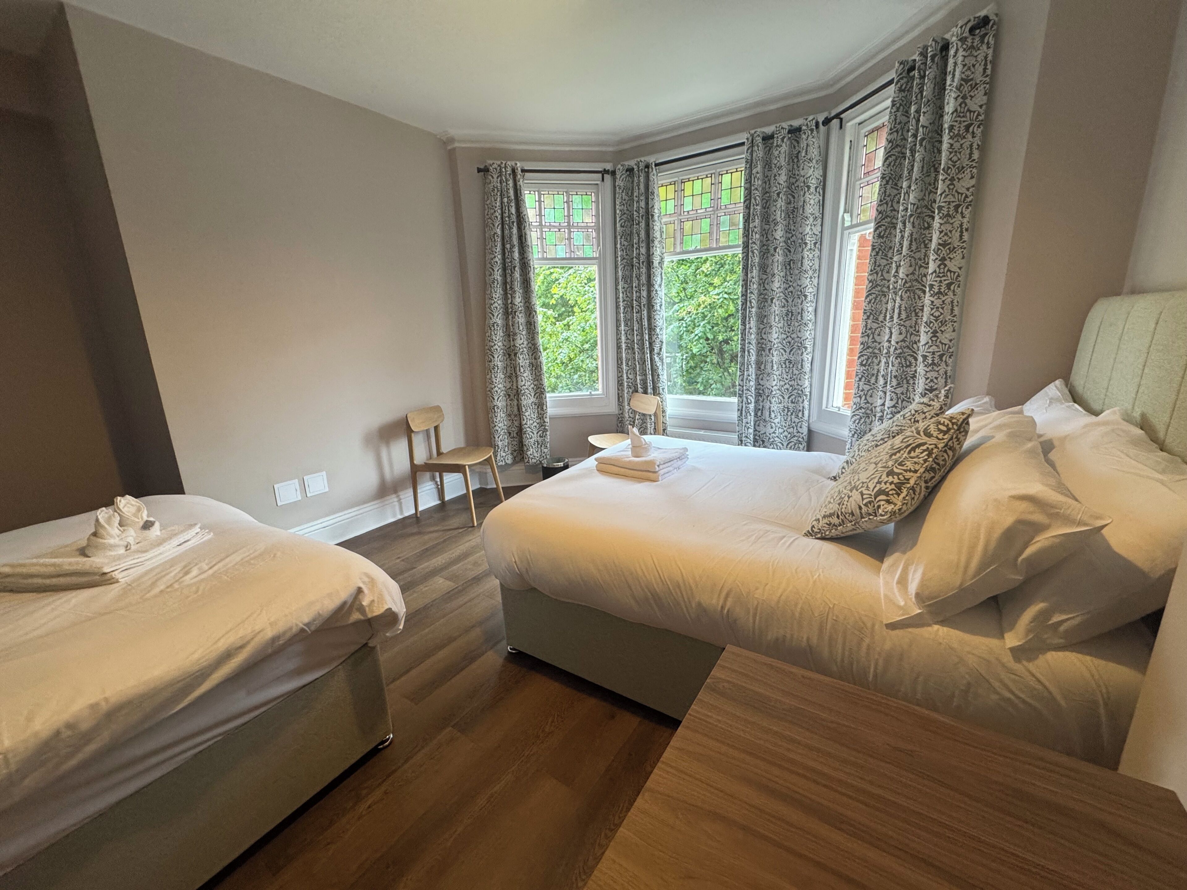 Een hotelkamer met twee bedden, een houten vloer, een stoel en ramen met gordijnen met een patroon.