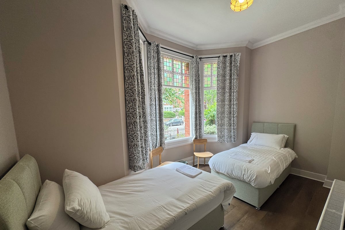 A bedroom with two beds, a chair, a radiator, and a window with curtains.