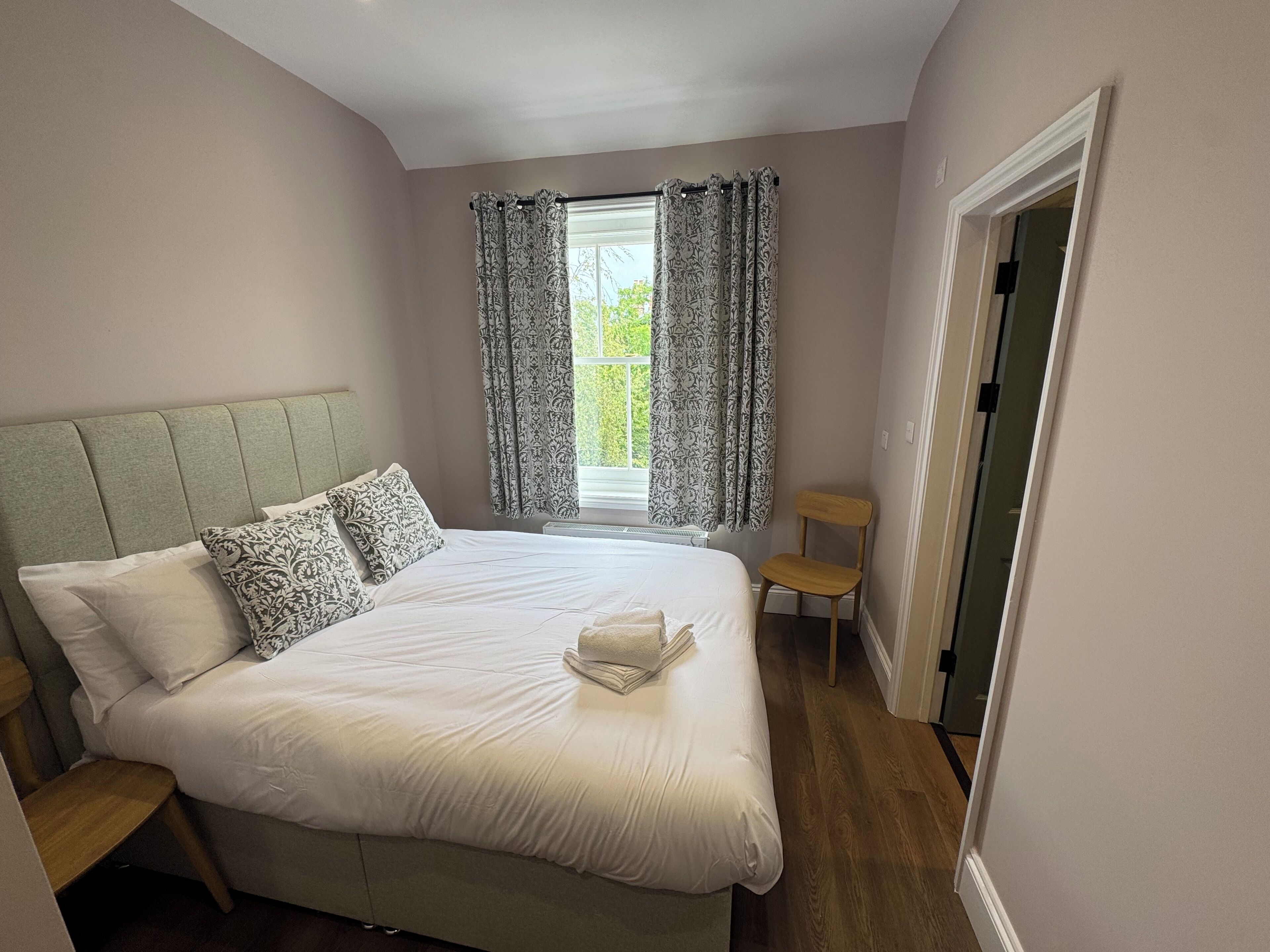 A neatly made bed with patterned pillows, a wooden chair, and a window with curtains.
