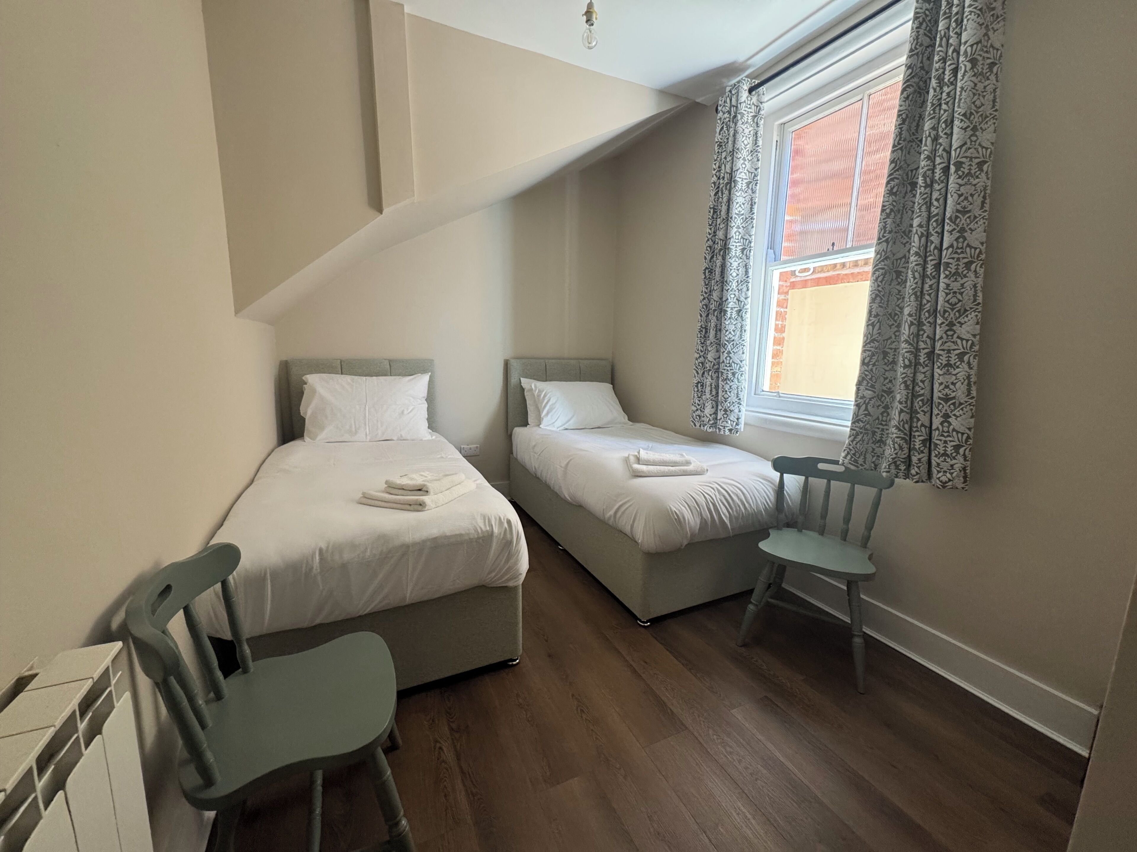 Un dormitorio con dos camas, una silla, un radiador y una ventana con cortinas.