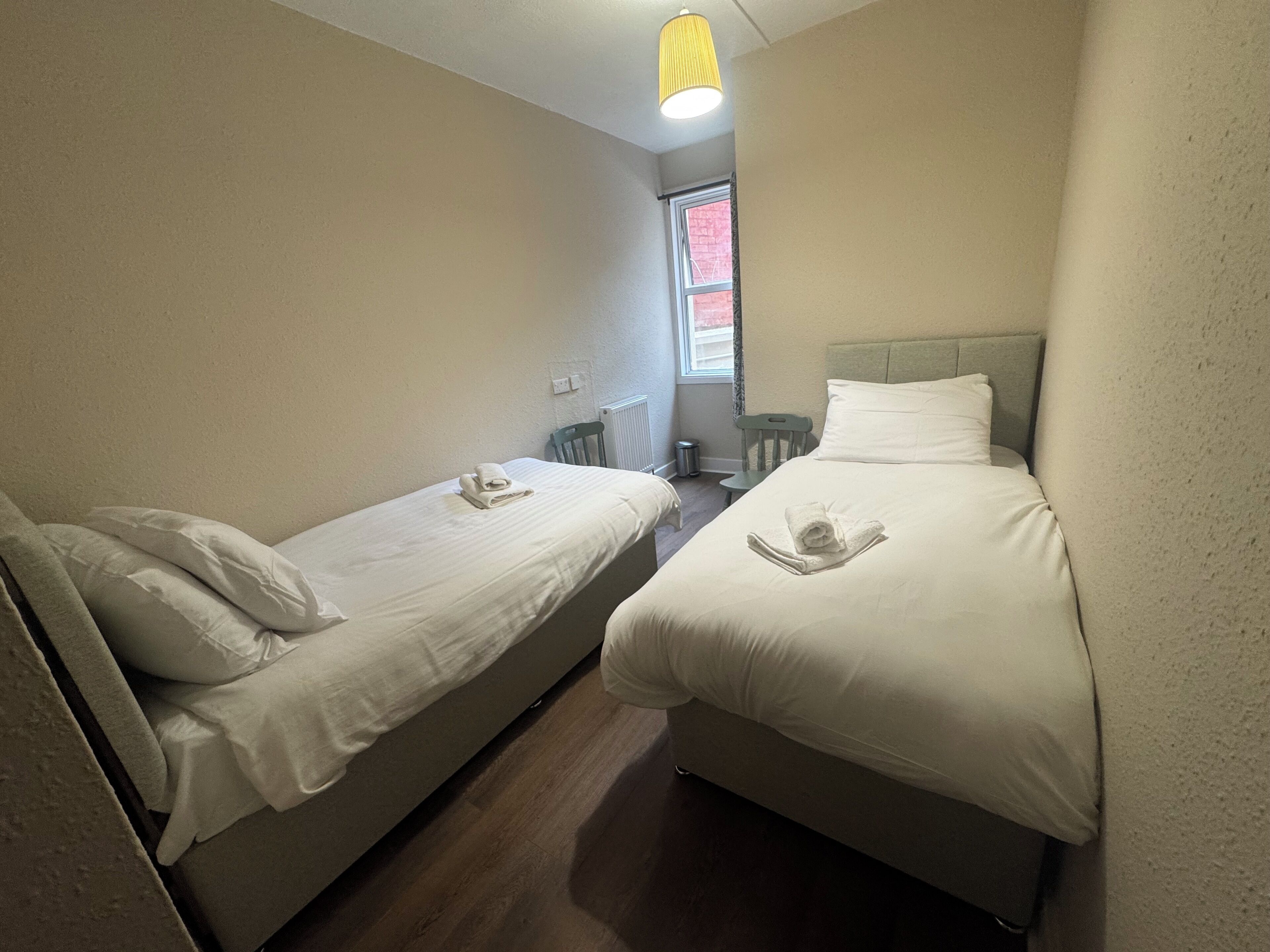 A room with two single beds, a small window, a radiator, and a chair.
