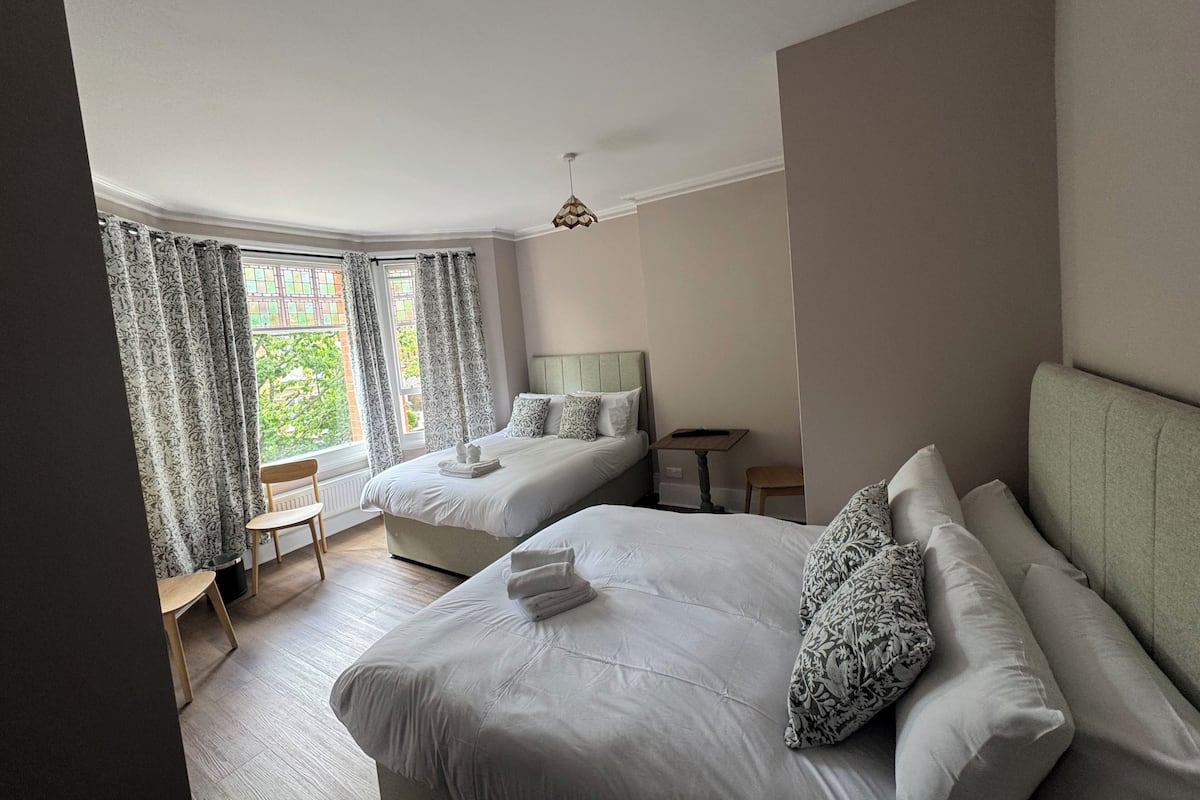 A hotel room with two beds, a small table, a chair, and a window with curtains.