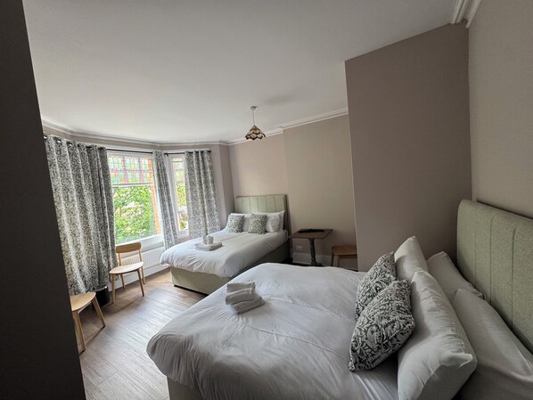 A hotel room with two beds, a small table, a chair, and a window with curtains.