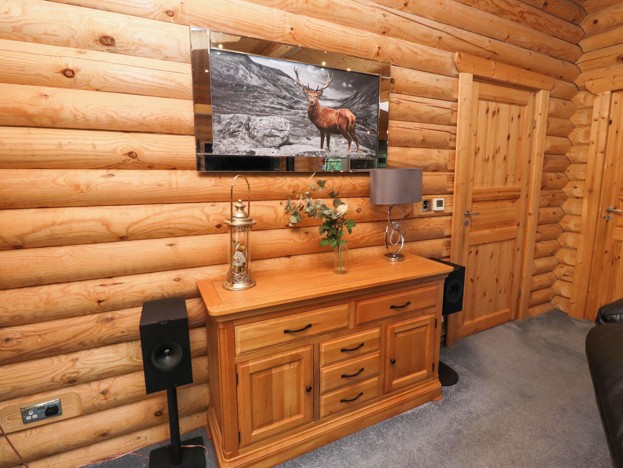 A wooden cabin interior with a mounted TV displaying a deer image, a wooden cabinet with drawers, a lamp, and a speaker.