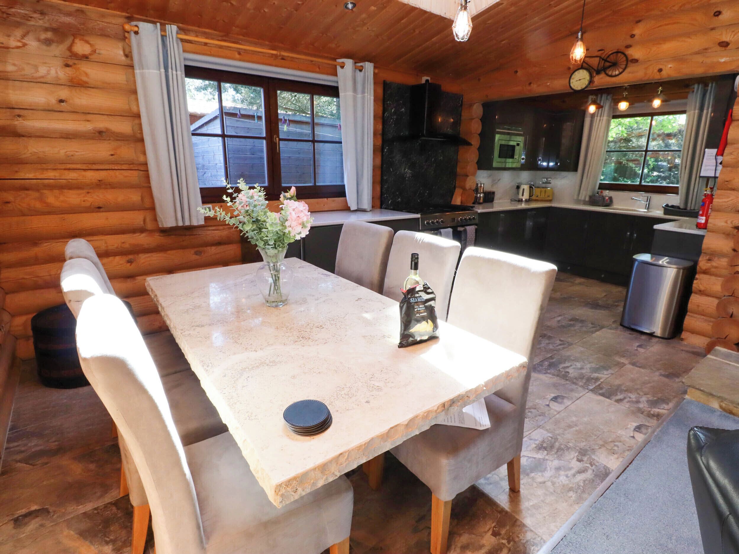 A wooden cabin interior with a dining table, chairs, a vase with flowers, a bag of snacks, and a kitchen area with a stove and sink.