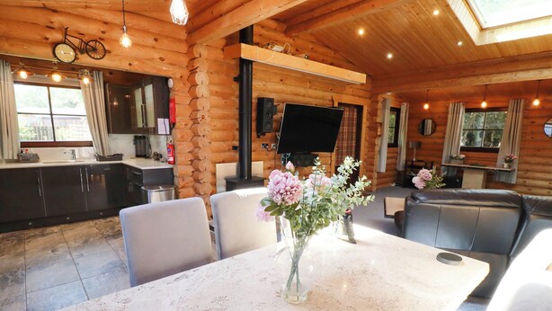 Interno di una baita di legno con tavolo da pranzo, sedie, divano, televisore e angolo cottura.