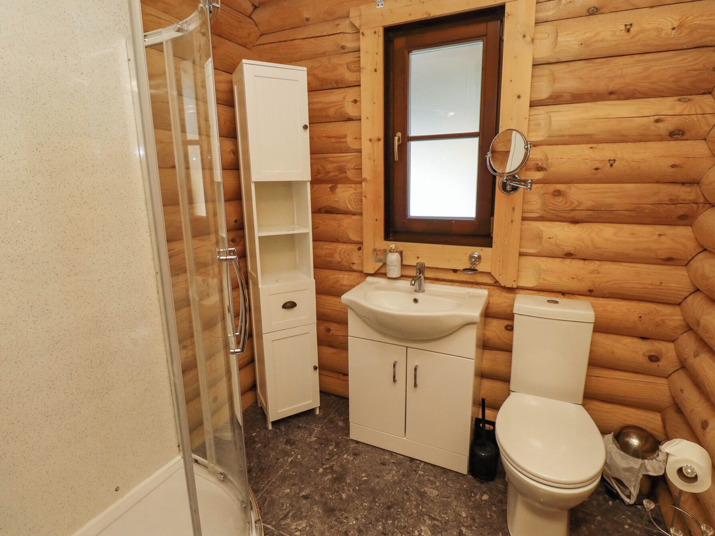 Een knus houten vakantiehuisje met een badkamer, inclusief douche, wastafel, toilet en een klein raam boven de wastafel.