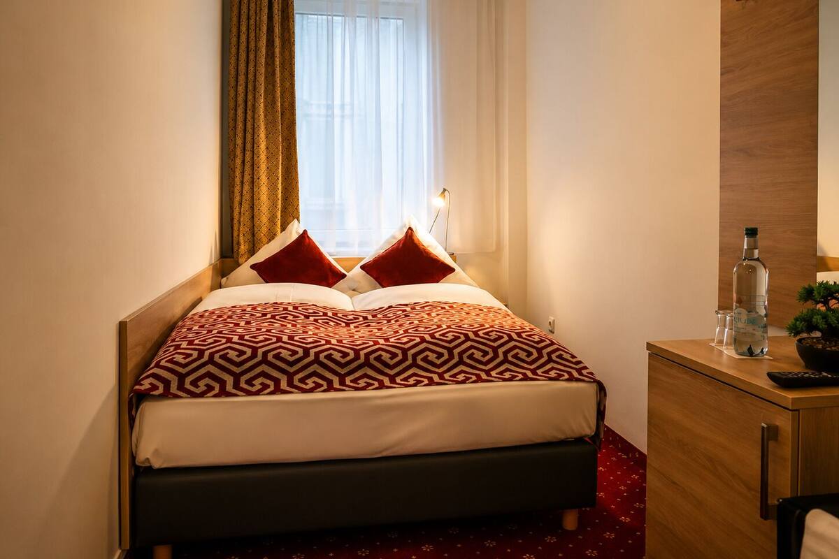 Ein Hotelzimmer mit einem Bett, einem Nachttisch mit Flasche und Gläsern, einem Holzschrank und einem Fenster mit Vorhängen.