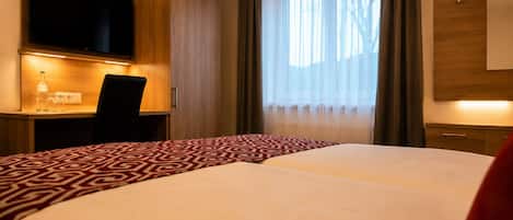 Habitación de hotel con cama, escritorio, televisión y ventana con cortinas.