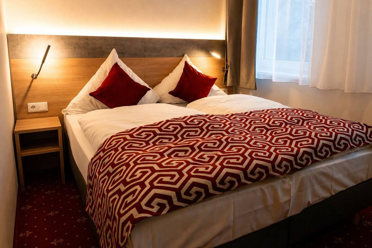 Ein Hotelzimmer mit Bett, zwei Kissen, Nachttisch, Lampe und einem Fenster mit Vorhängen.