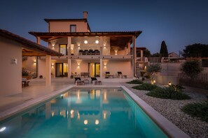 Una casa de dos pisos con piscina, mobiliario exterior y un patio bien iluminado.