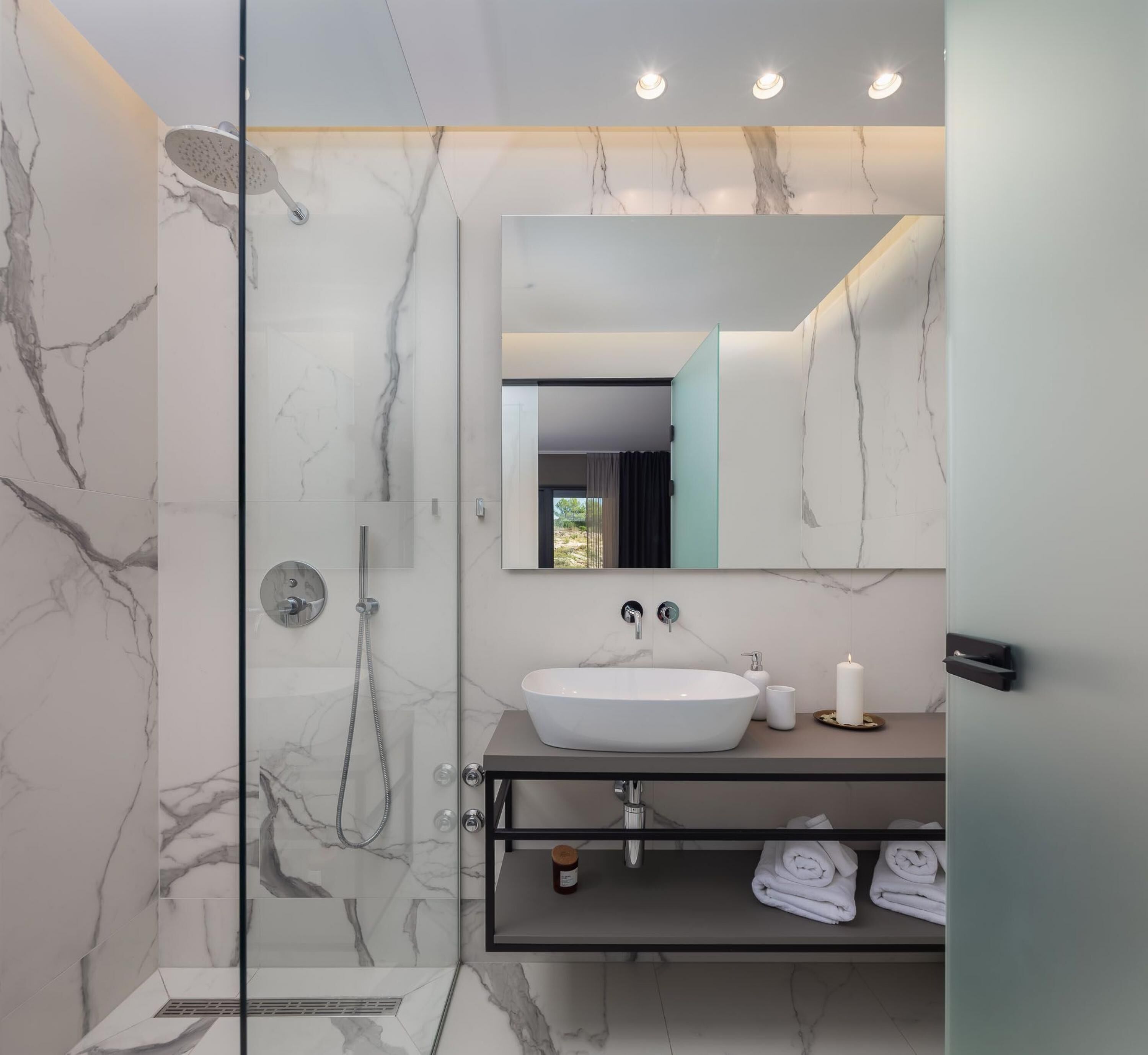 A modern bathroom with a glass-enclosed shower, a marble-tiled wall, a large mirror, and a vanity with a sink and towel rack.