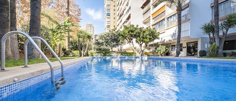 A swimming pool with metal railings, surrounded by palm trees and a multi-story building with balconies.