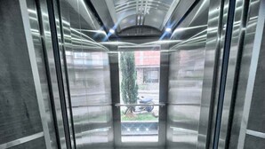 Een lift met metalen wanden en een glazen deur, leidend naar een buitenruimte met een scooter en groen.