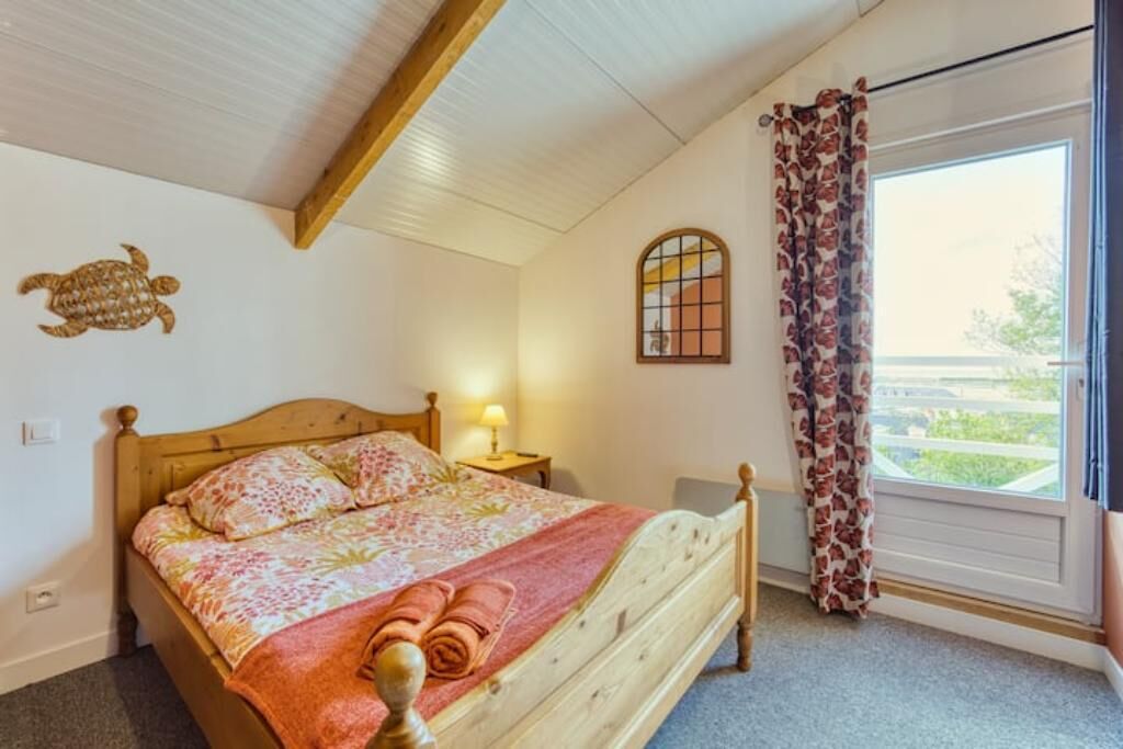 Een slaapkamer met een houten bed, bloemenbedrukking, een raam met gordijnen, een schildering van een schildpad aan de muur, en een klein nachtkastje met een lamp.