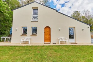 A single-story building with a beige exterior, a wooden front door, and two white plastic chairs in front. - Le Gîte de la Calotine 4 Étoiles (Dives-sur-Mer)
