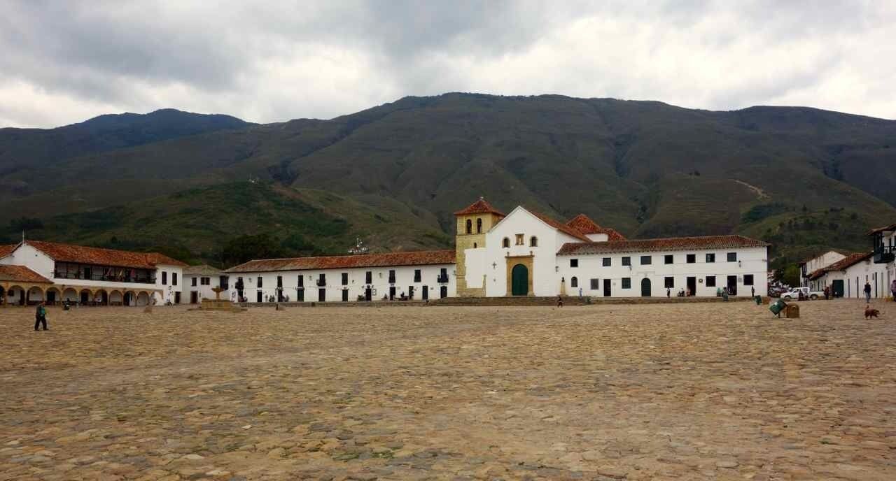Un patio con un edificio blanco que presenta una puerta verde y un campanario, rodeado de muros blancos y suelo adoquinado.