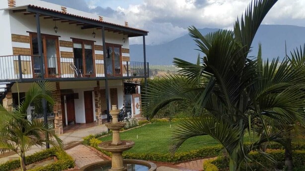 Un edificio de dos pisos con balcón, una fuente y un jardín bien cuidado con palmeras y arbustos.